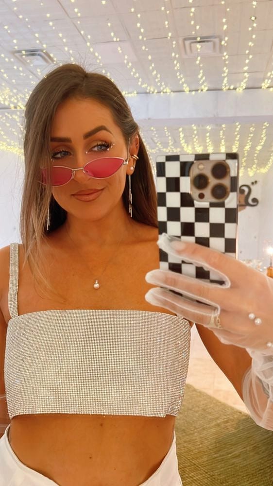A woman in sunglasses and a sparkly top taking a mirror selfie under string lights
